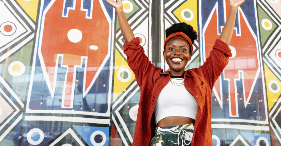 A lady stands infront of a wall painted with vibrant patterns, with her arms raised up and she is smiling.