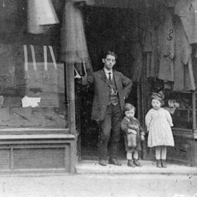 A black and white photograph showing an adult man and two young children standing in front of a clothing store.