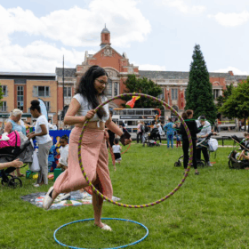 A woman stands in a grassy field with lots of other people around her. She is holding a hula hoop as if she's about to step into it.