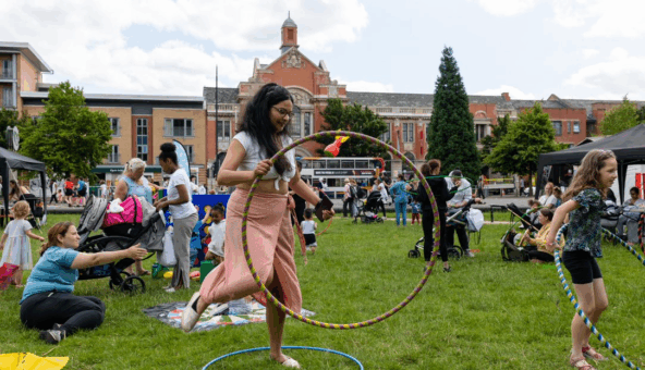A woman stands in a grassy field with lots of other people around her. She is holding a hula hoop as if she's about to step into it.