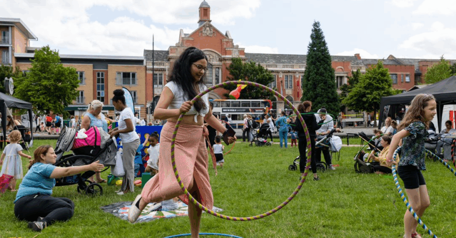 A woman stands in a grassy field with lots of other people around her. She is holding a hula hoop as if she's about to step into it.