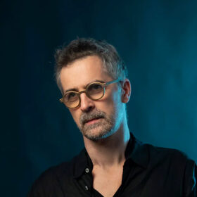 Sylvain Rifflet wearing round glasses stood on a deep blue background. The top of his saxophone reed is visible in the bottom right hand corner.