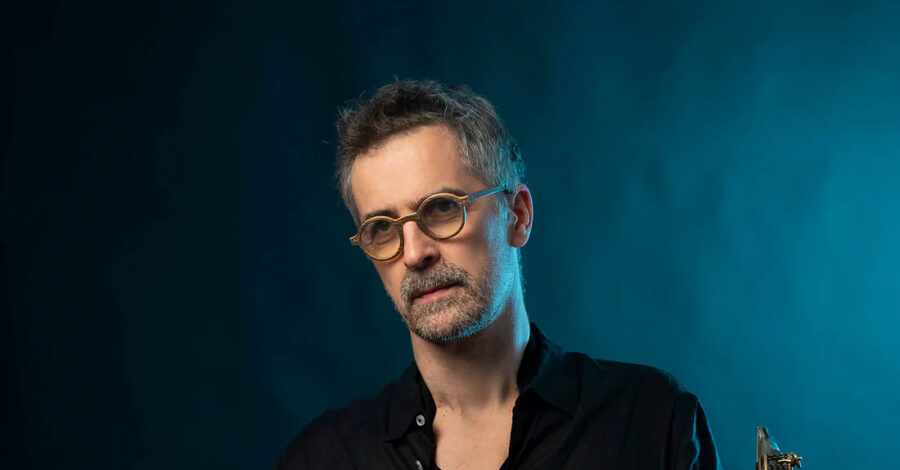 Sylvain Rifflet wearing round glasses stood on a deep blue background. The top of his saxophone reed is visible in the bottom right hand corner.