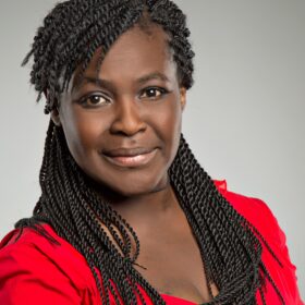 An image of Dame Maggie Aderin-Pocock, a black middle-aged woman with long hair in a red top, looking directly at the camera with a warm expression.