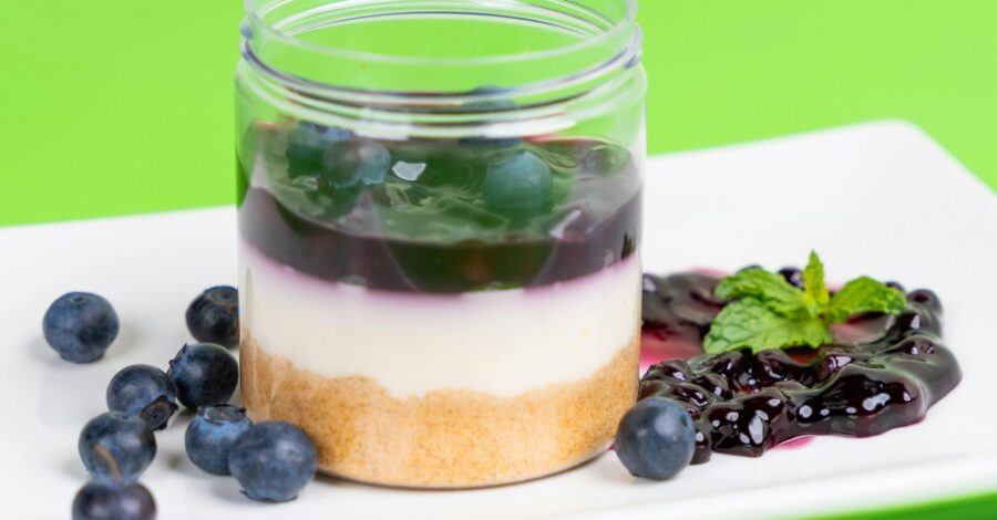A blueberry cheesecake in a glass jar container, on a white rectangular plate. There are some blueberries loose on the plate too.