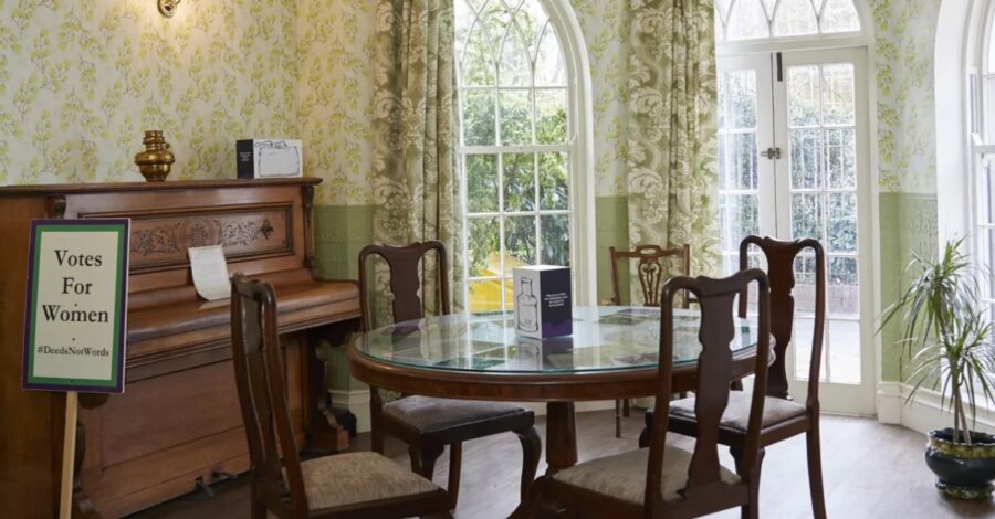 An Edwardian style room with piano, dining table and chairs, and a 'Votes for Women' placard