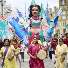 group of people dressed in bright colours and playing walking in front of large model of a woman also in bright colours