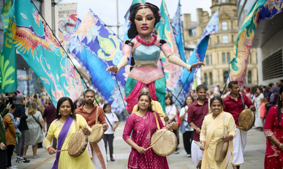 group of people dressed in bright colours and playing walking in front of large model of a woman also in bright colours
