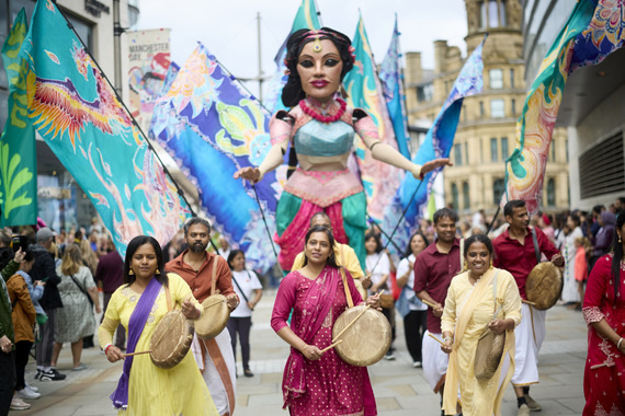 group of people dressed in bright colours and playing walking in front of large model of a woman also in bright colours