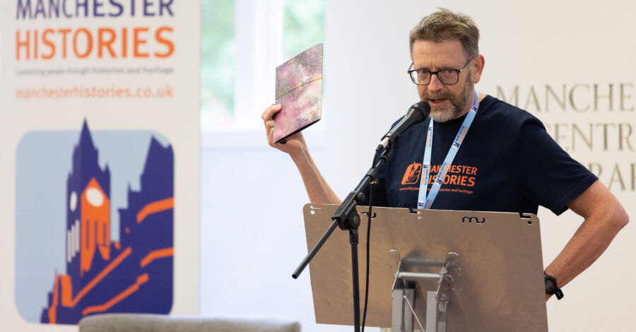 A volunteer at Manchester Histories Festival 2024 standing at a lectern holding a leaflet up.