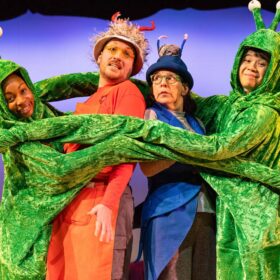 Four performers standing on stage with their arms around eachother. The Two performers on the outside are dressed in green, with long arms and antennas. There are two performers stood together in the middle, one dressed in orange and one dressed in blue.