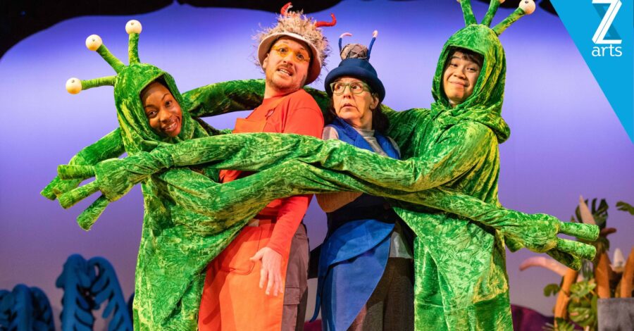 Four performers standing on stage with their arms around eachother. The Two performers on the outside are dressed in green, with long arms and antennas. There are two performers stood together in the middle, one dressed in orange and one dressed in blue.