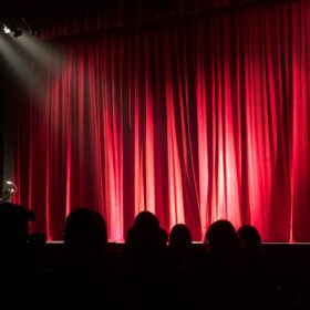 A lit stage with a red curtain closed across it. There are some heads and shoulders of the audience sat in seats in front of it as if they are waiting for the performance to start.