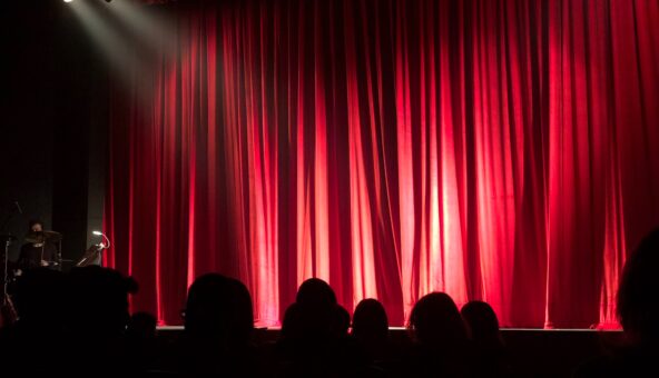 A lit stage with a red curtain closed across it. There are some heads and shoulders of the audience sat in seats in front of it as if they are waiting for the performance to start.