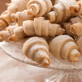 A close-up shot of a pile of freshly baked, crescent-shaped pastries, dusted generously with powdered sugar, resting on a clear glass cake stand. The background is softly blurred, highlighting the texture and inviting appearance of the pastries.