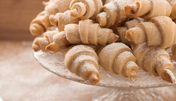 A close-up shot of a pile of freshly baked, crescent-shaped pastries, dusted generously with powdered sugar, resting on a clear glass cake stand. The background is softly blurred, highlighting the texture and inviting appearance of the pastries.