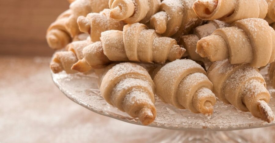 A close-up shot of a pile of freshly baked, crescent-shaped pastries, dusted generously with powdered sugar, resting on a clear glass cake stand. The background is softly blurred, highlighting the texture and inviting appearance of the pastries.