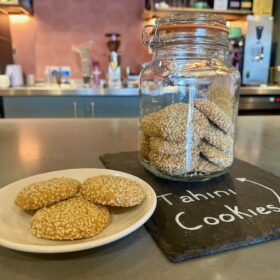 A still life image displays a white plate with three round, golden-brown cookies heavily coated in sesame seeds, positioned next to a large glass jar filled with more of the same cookies. The jar sits on a dark slate board with "Tahini Cookies" written in chalk. The background shows a blurred cafe counter with various equipment.