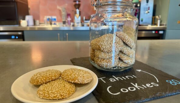 A still life image displays a white plate with three round, golden-brown cookies heavily coated in sesame seeds, positioned next to a large glass jar filled with more of the same cookies. The jar sits on a dark slate board with "Tahini Cookies" written in chalk. The background shows a blurred cafe counter with various equipment.