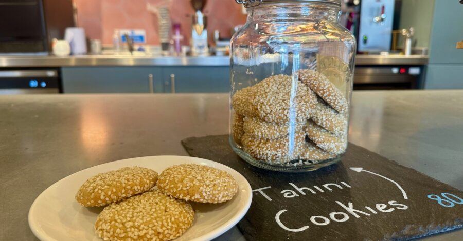 A still life image displays a white plate with three round, golden-brown cookies heavily coated in sesame seeds, positioned next to a large glass jar filled with more of the same cookies. The jar sits on a dark slate board with "Tahini Cookies" written in chalk. The background shows a blurred cafe counter with various equipment.