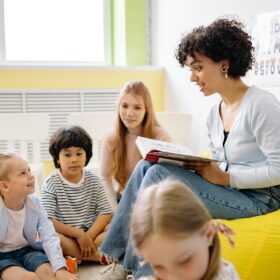 A woman sat on a yellow beanbag holding a book, reading a story to a group of children who are sat on the floor.