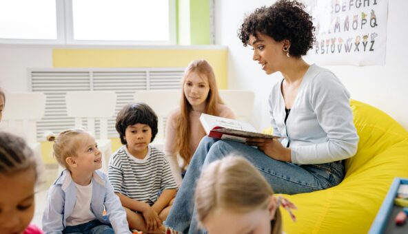 A woman sat on a yellow beanbag holding a book, reading a story to a group of children who are sat on the floor.