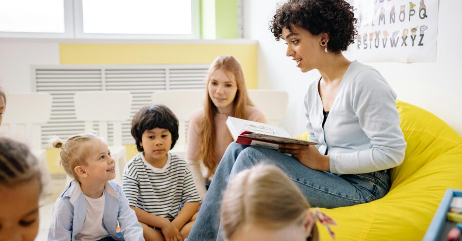 A woman sat on a yellow beanbag holding a book, reading a story to a group of children who are sat on the floor.