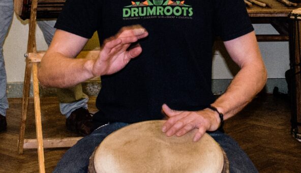 A person wearing a black 'Drumroots' t-shirt playing a drum.