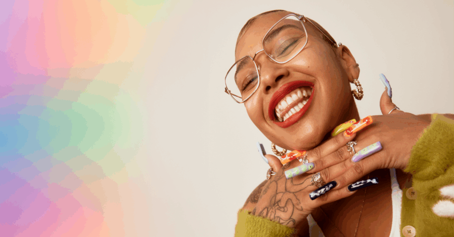 The artist Jazmin Morris smiling with her hands interlinked below her neck. She stands in front of a rainbow coloured background.