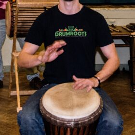 A person wearing a black 'Drumroots' t-shirt playing a drum.