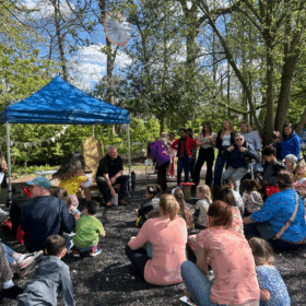 A blue gazebo and a number of families and people sat on the floor and stood up ready for a storytelling event.