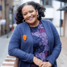 Poet Lorna Goodison smiling and leaning on a brick wall outside.