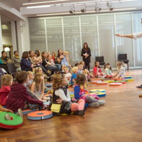 An audience of young people sat on a wooden floor in a hall watching a performer who is standing up in front of them.