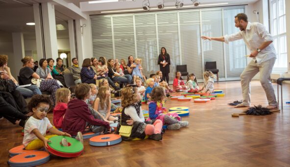 An audience of young people sat on a wooden floor in a hall watching a performer who is standing up in front of them.