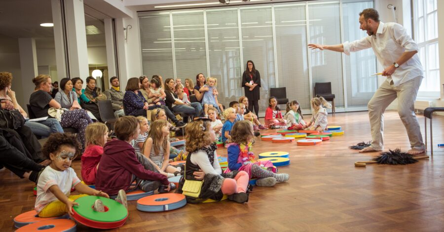 An audience of young people sat on a wooden floor in a hall watching a performer who is standing up in front of them.