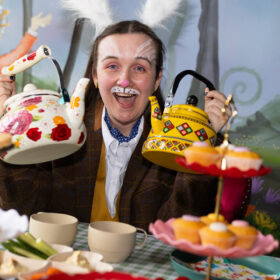 A woman dressed as the white rabbit holds up two brightly coloured teapots to the camera.