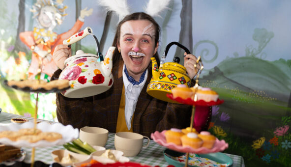 A woman dressed as the white rabbit holds up two brightly coloured teapots to the camera.