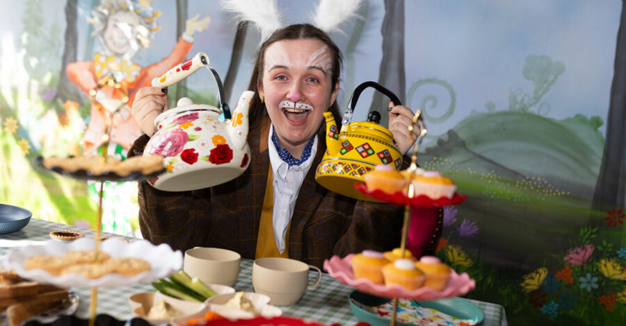 A woman dressed as the white rabbit holds up two brightly coloured teapots to the camera.