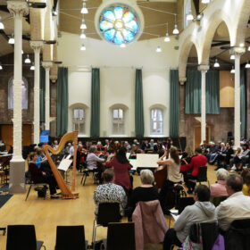 A stage with a musician playing a harp and an audience sat on chairs watching inside Halle St Peters.