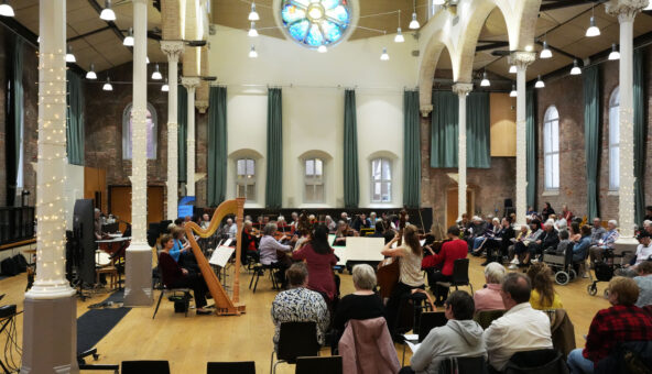A stage with a musician playing a harp and an audience sat on chairs watching inside Halle St Peters.