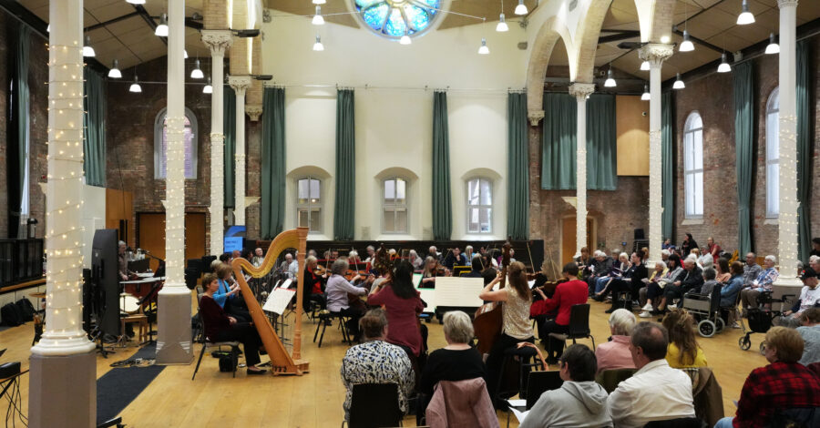 A stage with a musician playing a harp and an audience sat on chairs watching inside Halle St Peters.