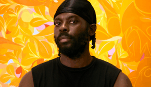 The artist Tunji Adeniyi-Jones wearing a black vest and a durag, stood in front of one of his artworks.