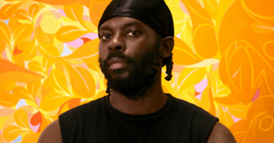 The artist Tunji Adeniyi-Jones wearing a black vest and a durag, stood in front of one of his artworks.