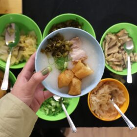 A hand holding a blue bowl of rice porridge topped with fried dough sticks, pork, pickled vegetables, and green onions. Below, several green and yellow bowls filled with different toppings and condiments, such as mushrooms, cabbage, boiled egg, and fried dough, are laid out with serving spoons.