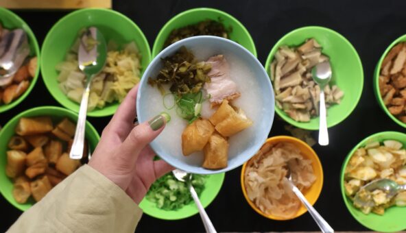 A hand holding a blue bowl of rice porridge topped with fried dough sticks, pork, pickled vegetables, and green onions. Below, several green and yellow bowls filled with different toppings and condiments, such as mushrooms, cabbage, boiled egg, and fried dough, are laid out with serving spoons.