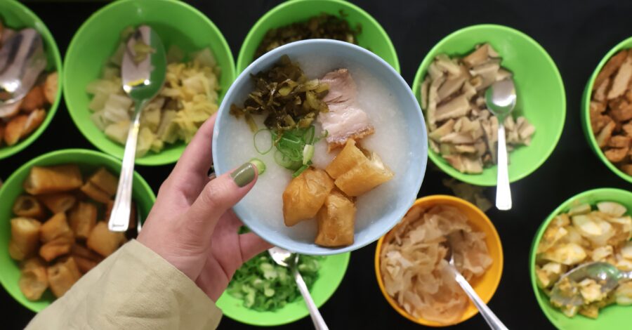 A hand holding a blue bowl of rice porridge topped with fried dough sticks, pork, pickled vegetables, and green onions. Below, several green and yellow bowls filled with different toppings and condiments, such as mushrooms, cabbage, boiled egg, and fried dough, are laid out with serving spoons.