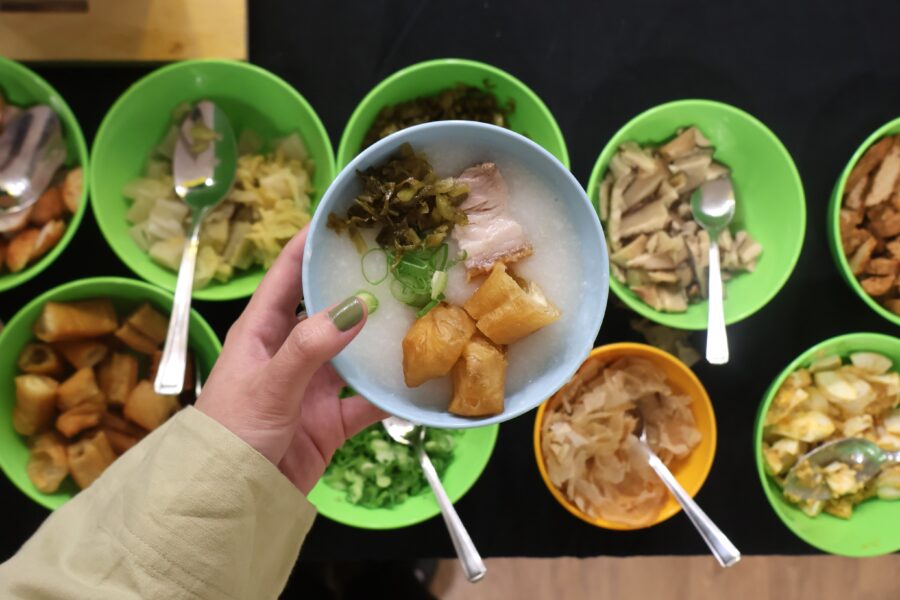 A hand holding a blue bowl of rice porridge topped with fried dough sticks, pork, pickled vegetables, and green onions. Below, several green and yellow bowls filled with different toppings and condiments, such as mushrooms, cabbage, boiled egg, and fried dough, are laid out with serving spoons.