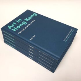 A stack of six hardcover books with dark blue covers titled Art in Hong Kong: Portrait of a City in Flux by Enid Tsui. The text on the cover and spine is printed in teal and white. The books are neatly stacked on a light-colored surface.
