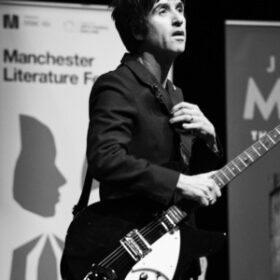 johnny marr wearing a dark suit wih a rickenbacker guitar in front of a poster for manchester literature festival