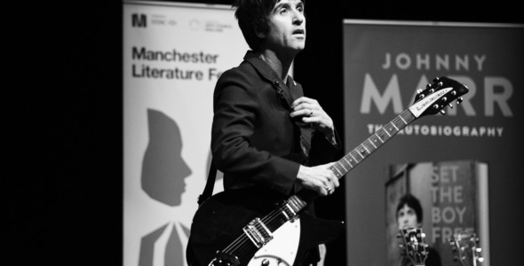 johnny marr wearing a dark suit wih a rickenbacker guitar in front of a poster for manchester literature festival